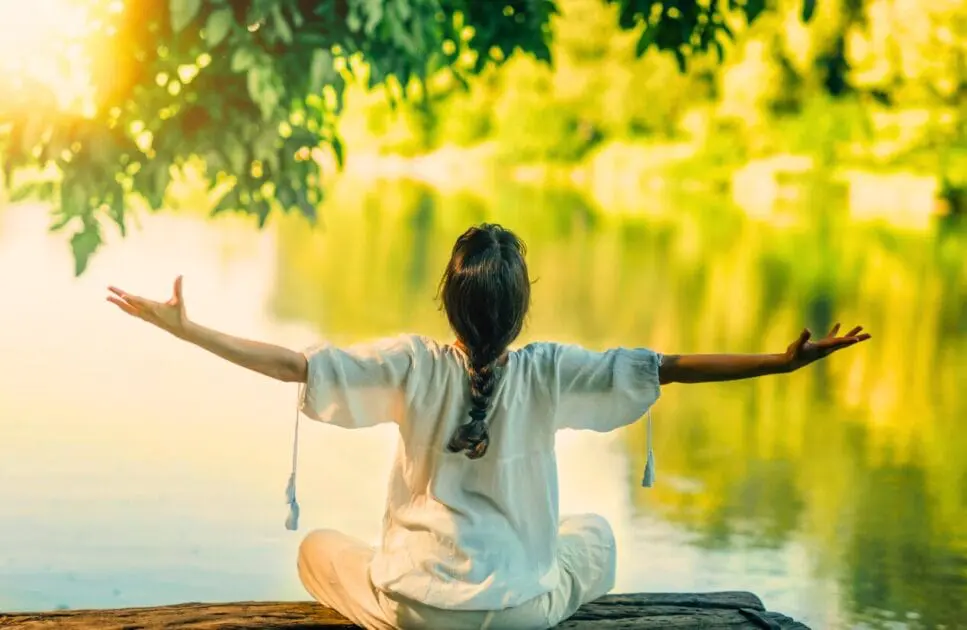 Woman sitting on a log by the river with her arms outstretched, embracing the serene natural surroundings. Hypnotheray near Geebung. Brisbane Livewell Clinic
